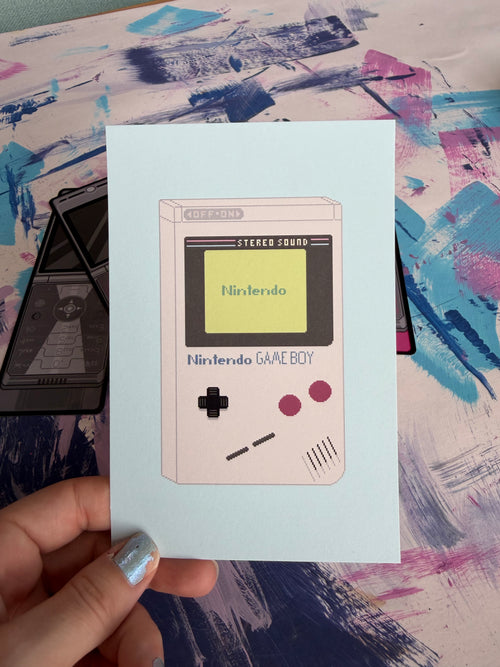 A hand with glittery silver nail polish holds a square art print featuring a retro Game Boy illustration on a light blue background. The print is placed over a stack of black Game Boy cases on a colorful, paint-splattered surface.