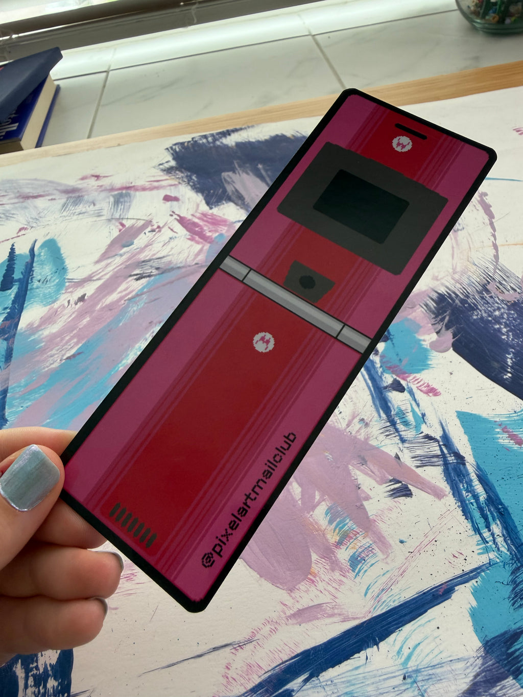 A hand with blue nail polish holds a custom-printed flip bookmark featuring a red and pink pixel art design, set against a colorful abstract painted background.