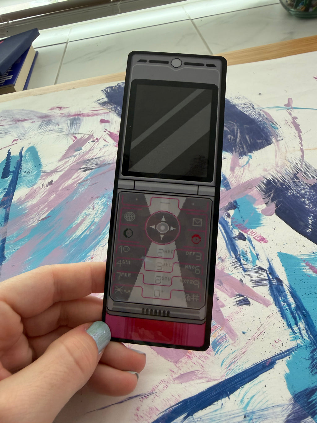 A black flip phone bookmark with a screen on the left and a keypad on the right, held in a hand against a colorful abstract painted background.