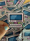 A hand with light blue glitter nail polish holds a holographic sticker shaped like a retro computer monitor. The sticker features a pink border, a dark blue screen with light blue lines, and a black-and-white pixelated keyboard base.