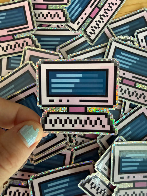 A hand with light blue glitter nail polish holds a holographic sticker shaped like a retro computer monitor. The sticker features a pink border, a dark blue screen with light blue lines, and a black-and-white pixelated keyboard base.